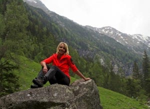 Frau mit roter Jacke sitzt auf Stein mit Gebirge im Hintergrund