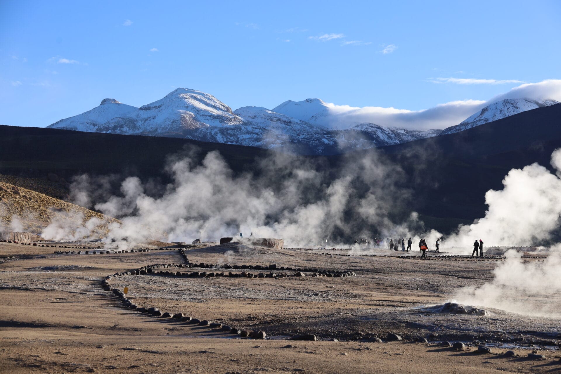 Die Menschen spazieren zwischen dampfenden Geysiren mit schneebedeckten Bergen im Hintergrund unter einem klaren blauen Himmel.