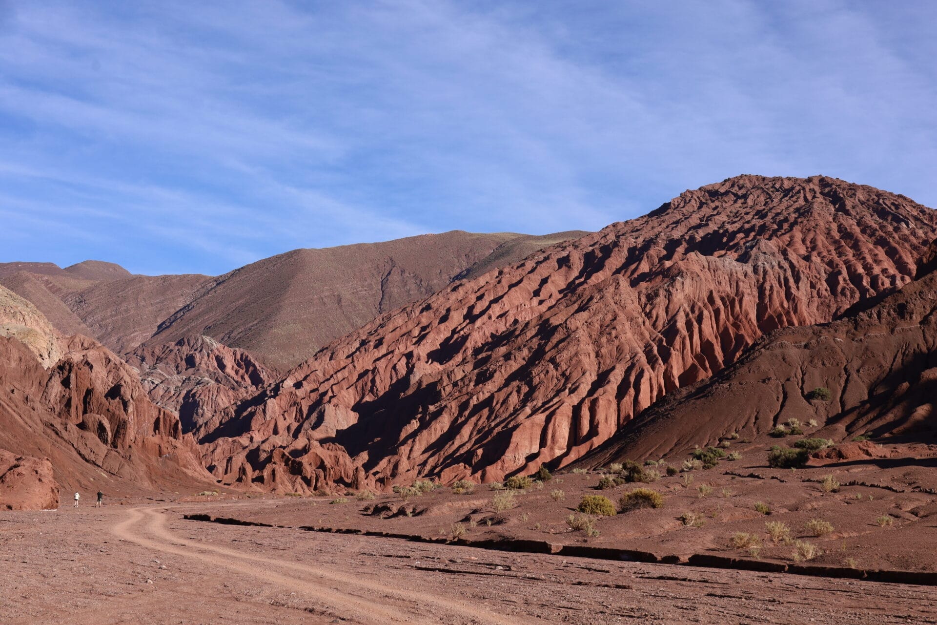 Rote und braune felsige Hügel mit zerklüfteten Strukturen erheben sich neben einer unbefestigten Straße unter einem teilweise bewölkten blauen Himmel in einer Wüstenlandschaft.