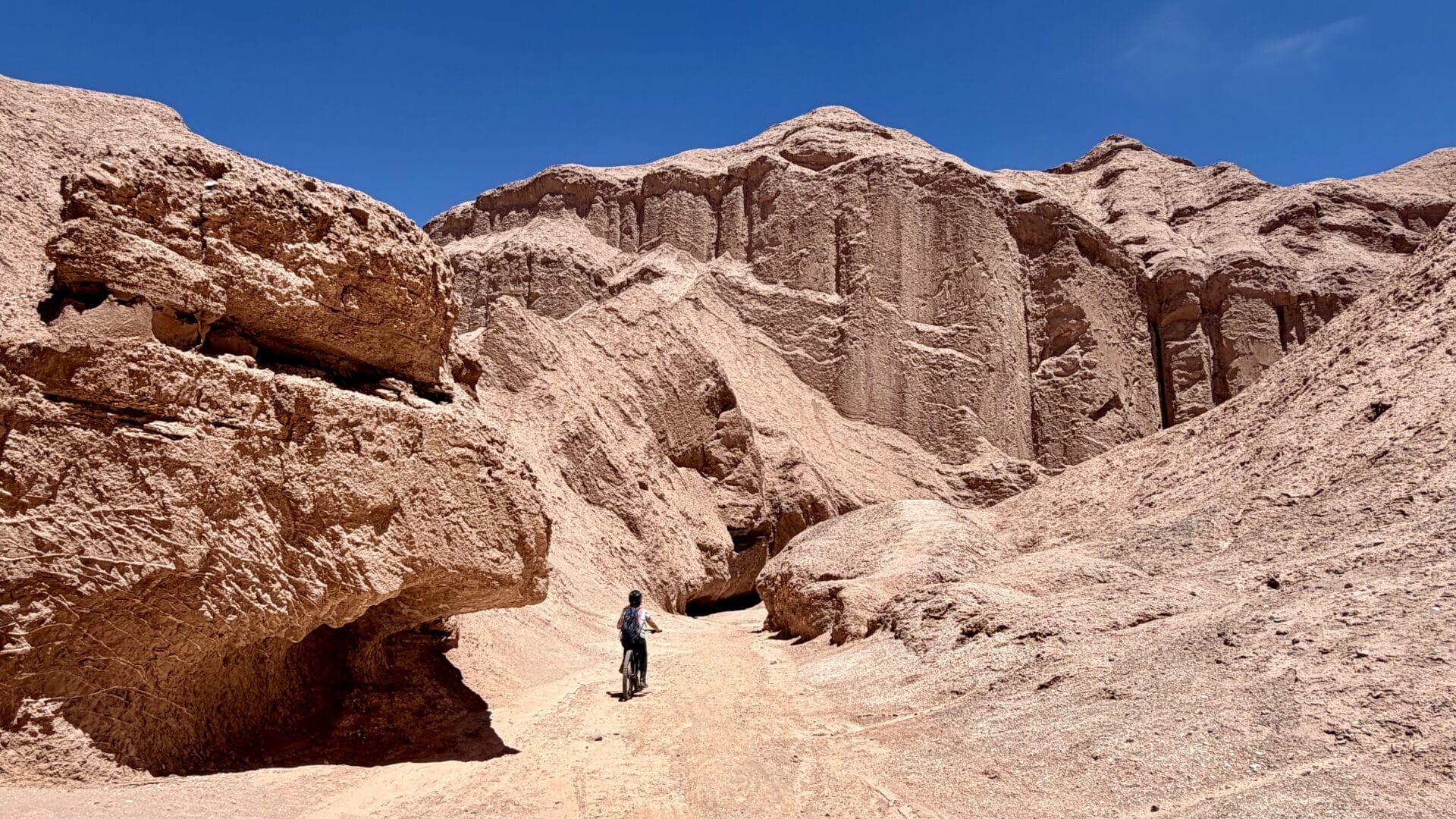Eine Person fährt mit dem Fahrrad in der chilenischen Atacama-Wüste, umgeben von großen, zerklüfteten Felsformationen unter einem klaren blauen Himmel.