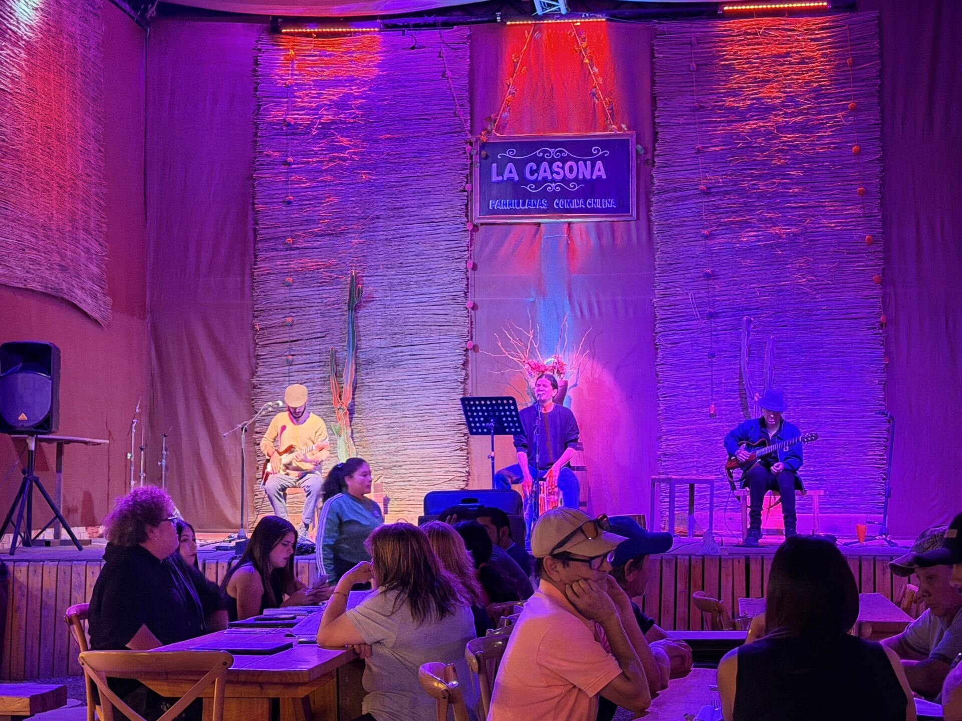 In einem Restaurant namens La Casona spielt eine kleine Band live auf der Bühne und erweckt den pulsierenden Geist der chilenischen Atacama-Wüste zum Leben, während die Menschen an den Tischen sitzen und bei bunter Beleuchtung essen und sich unterhalten.