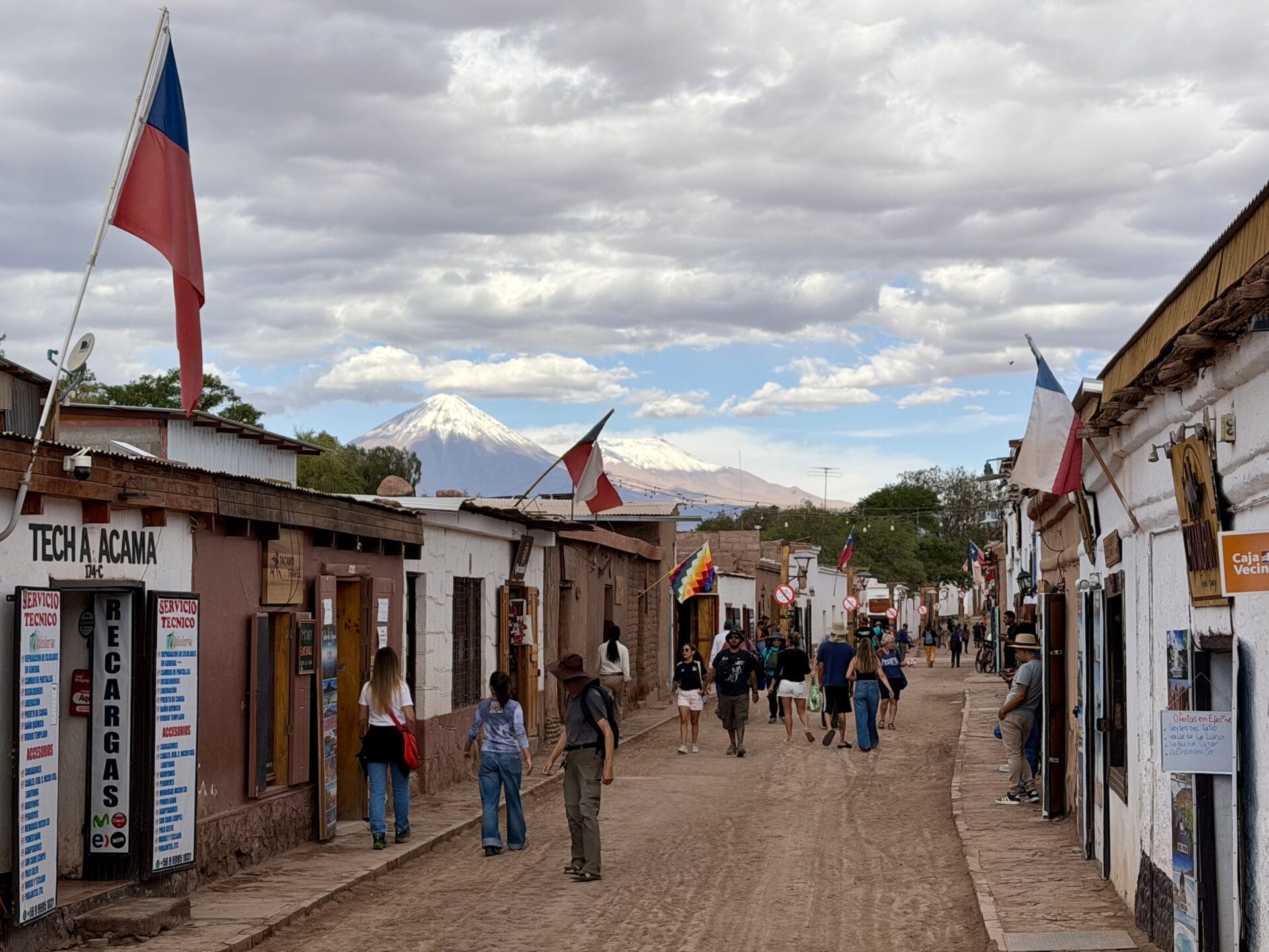 Menschen gehen eine unbefestigte Straße entlang, die von Geschäften und Gebäuden mit chilenischen Flaggen in der Atacama-Wüste Chiles gesäumt ist. Im Hintergrund ist ein schneebedeckter Berg unter einem bewölkten Himmel zu sehen.