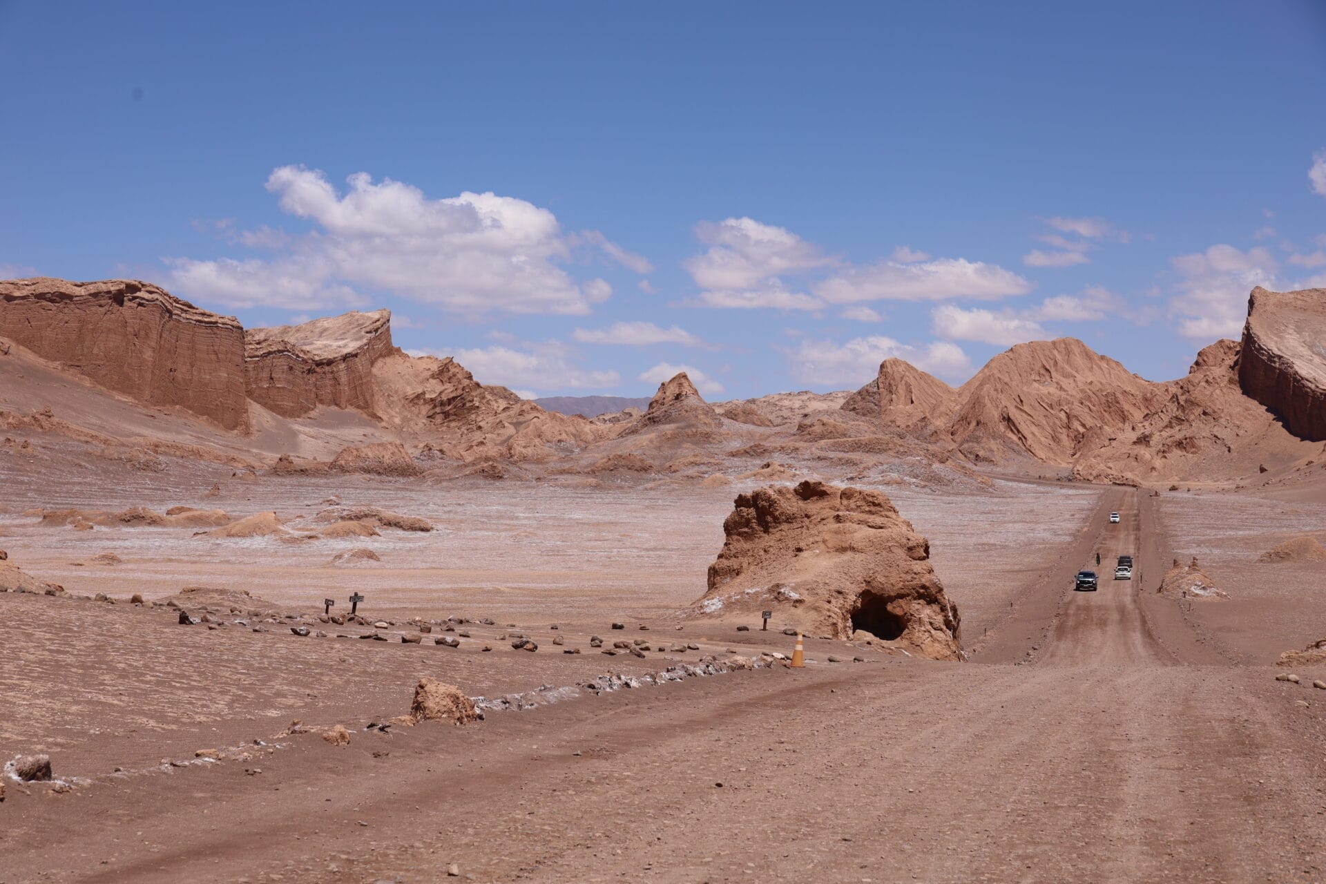 Eine asphaltierte Straße windet sich durch die karge Atacama-Wüste Chiles, flankiert von Felsformationen und fernen Bergen unter einem blauen Himmel mit vereinzelten Wolken. Mehrere Autos fahren auf dieser abgelegenen Wüstenroute.