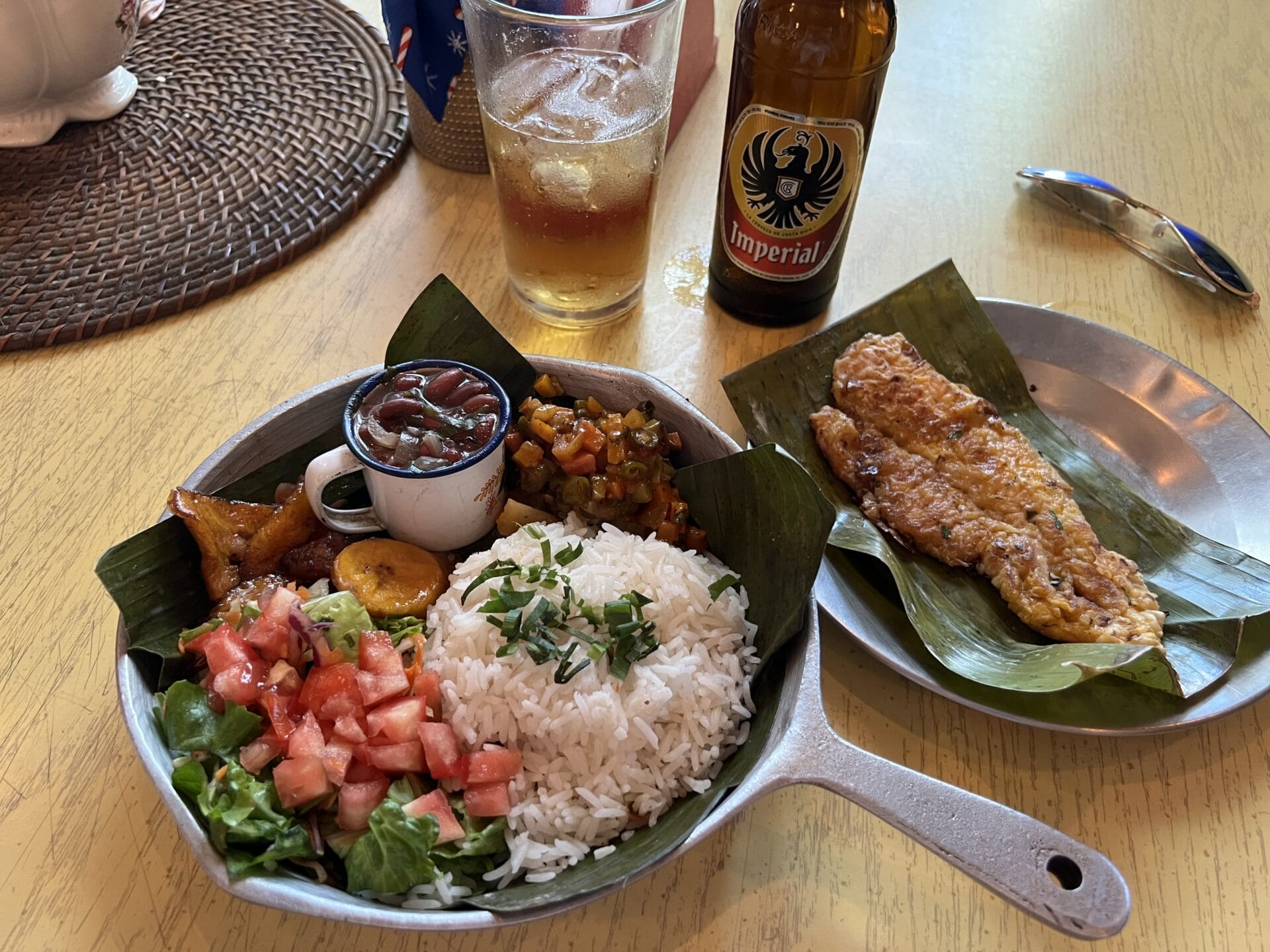 Eine traditionelle costa-ricanische Mahlzeit mit Reis, Bohnen, Salat, frittierten Kochbananen und Fisch, serviert auf einem Metallteller, dazu eine Flasche Imperial-Bier.