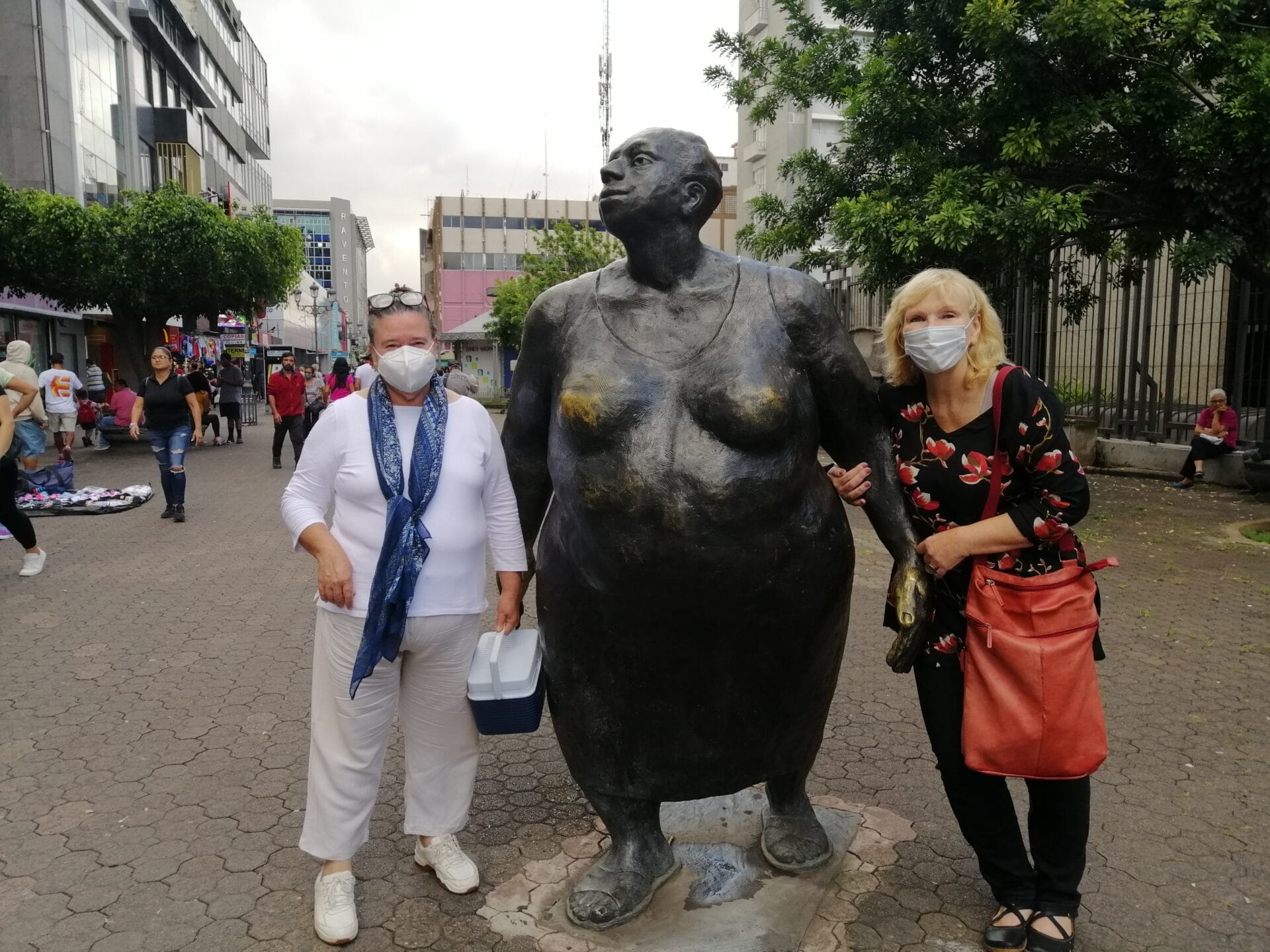 Zwei Frauen mit Masken stehen neben der Bronzestatue einer stehenden Figur auf einem städtischen Platz.