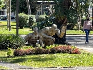 Eine Bronzestatue zweier älterer Menschen, einer sitzend, der andere hockend, aufgestellt in einem üppigen Park mit Passanten und Grün im Hintergrund.