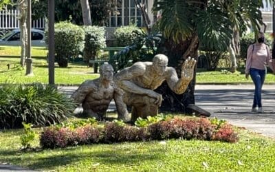 Eine Bronzestatue zweier älterer Menschen, einer sitzend, der andere hockend, aufgestellt in einem üppigen Park mit Passanten und Grün im Hintergrund.