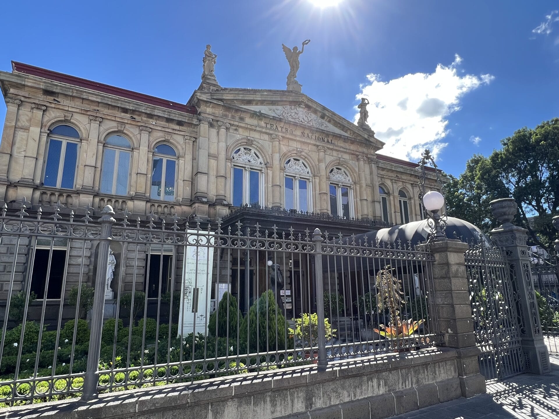 Fassade eines neoklassizistischen Gebäudes mit dekorativen Skulpturen und dahinter scheinender Sonne, umgeben von einem verzierten Metallzaun, unter einem klaren blauen Himmel.