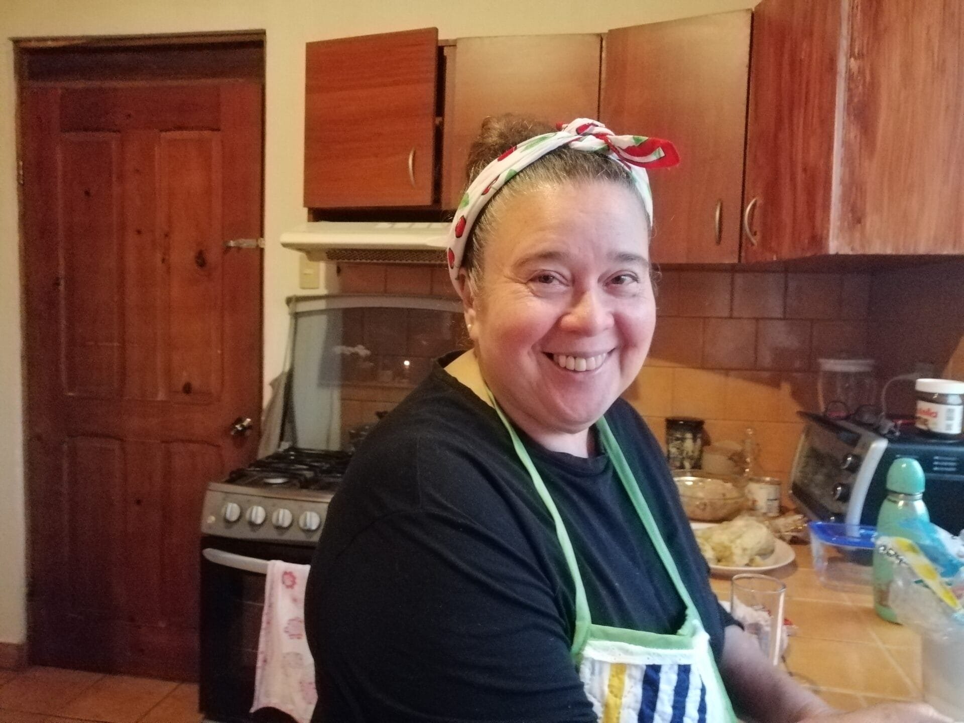 Eine lächelnde Frau mit Stirnband und Schürze steht in einer Küche mit Holzschränken und einem Herd im Hintergrund.