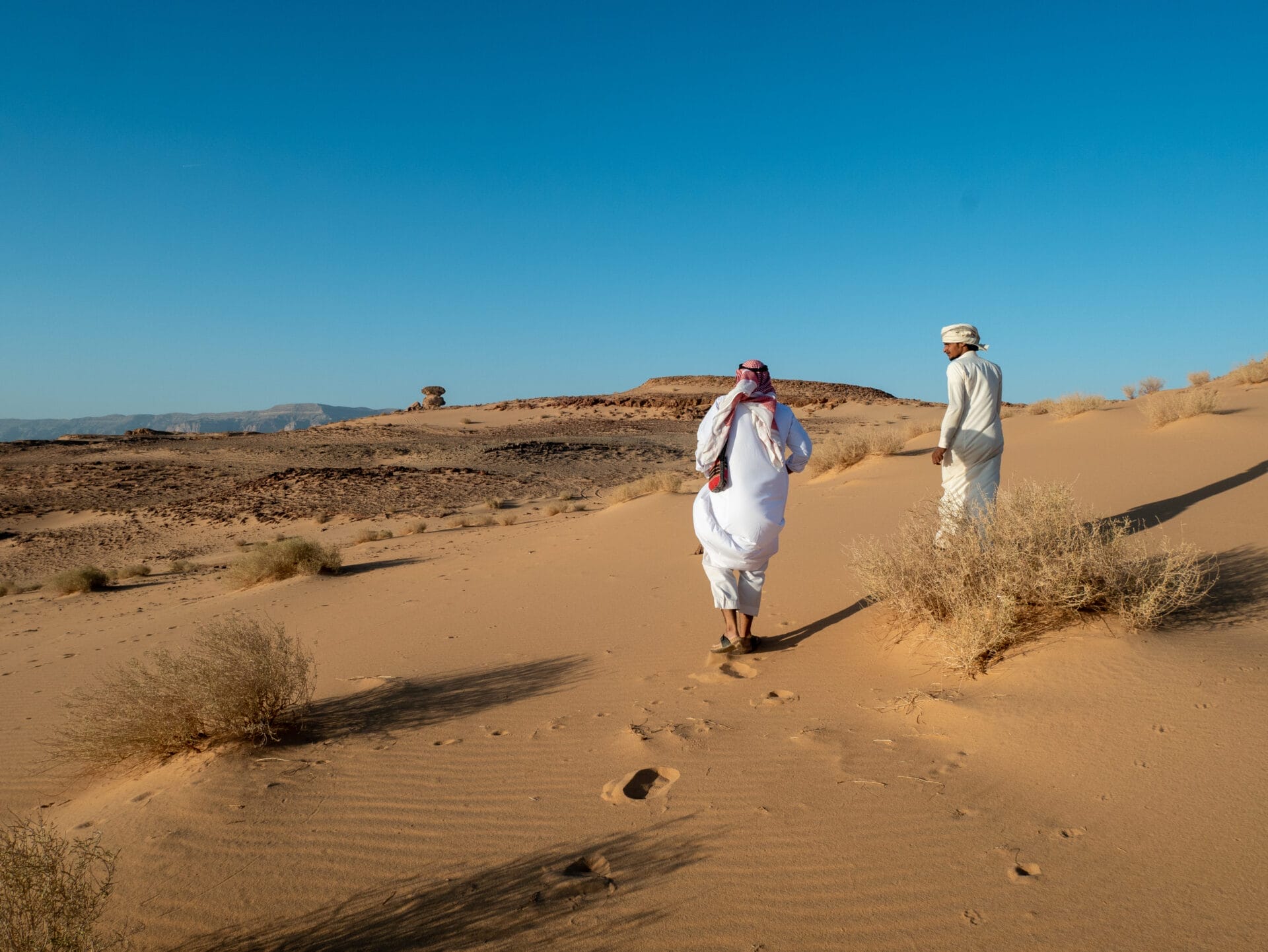 Zwei Menschen in traditioneller Kleidung laufen unter einem klaren blauen Himmel durch eine sandige Wüstenlandschaft.