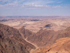 Ein Panoramablick auf eine weite, trockene Wüstenlandschaft mit schroffen, felsigen Bergen unter einem blauen Himmel mit dünnen Wolken.