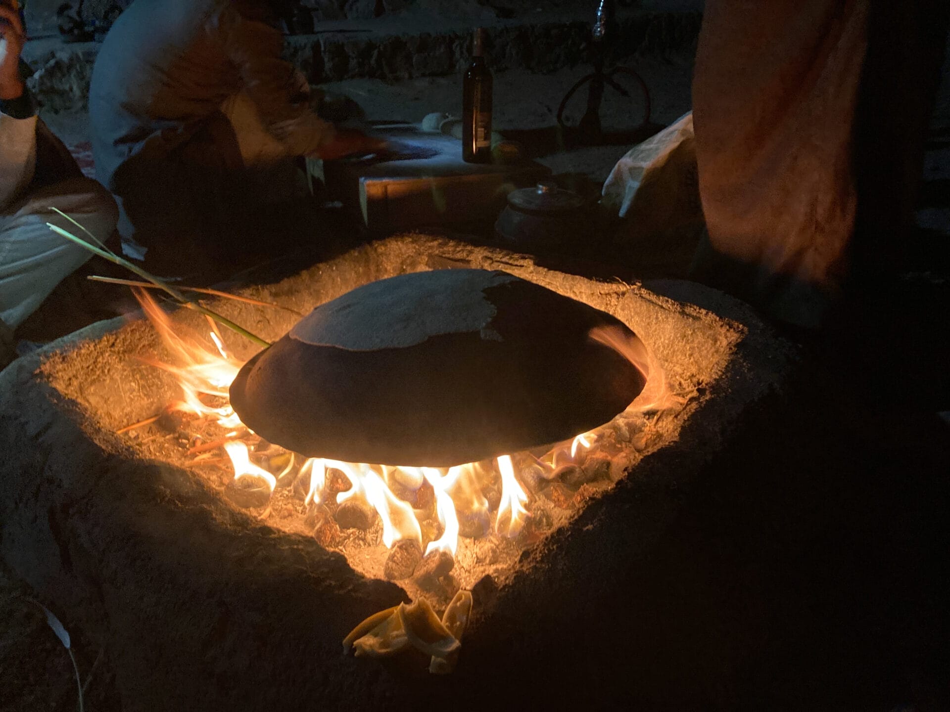 Eine kuppelförmige Metallpfanne wird über einer offenen Flamme am Lagerfeuer erhitzt, während Menschen um sie herum sitzen.