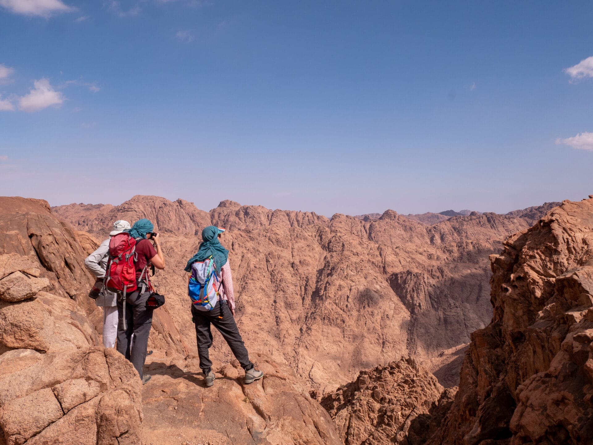 Drei Wanderer mit Rucksäcken stehen auf einer Felskante und blicken auf eine weite Berglandschaft unter einem klaren blauen Himmel.