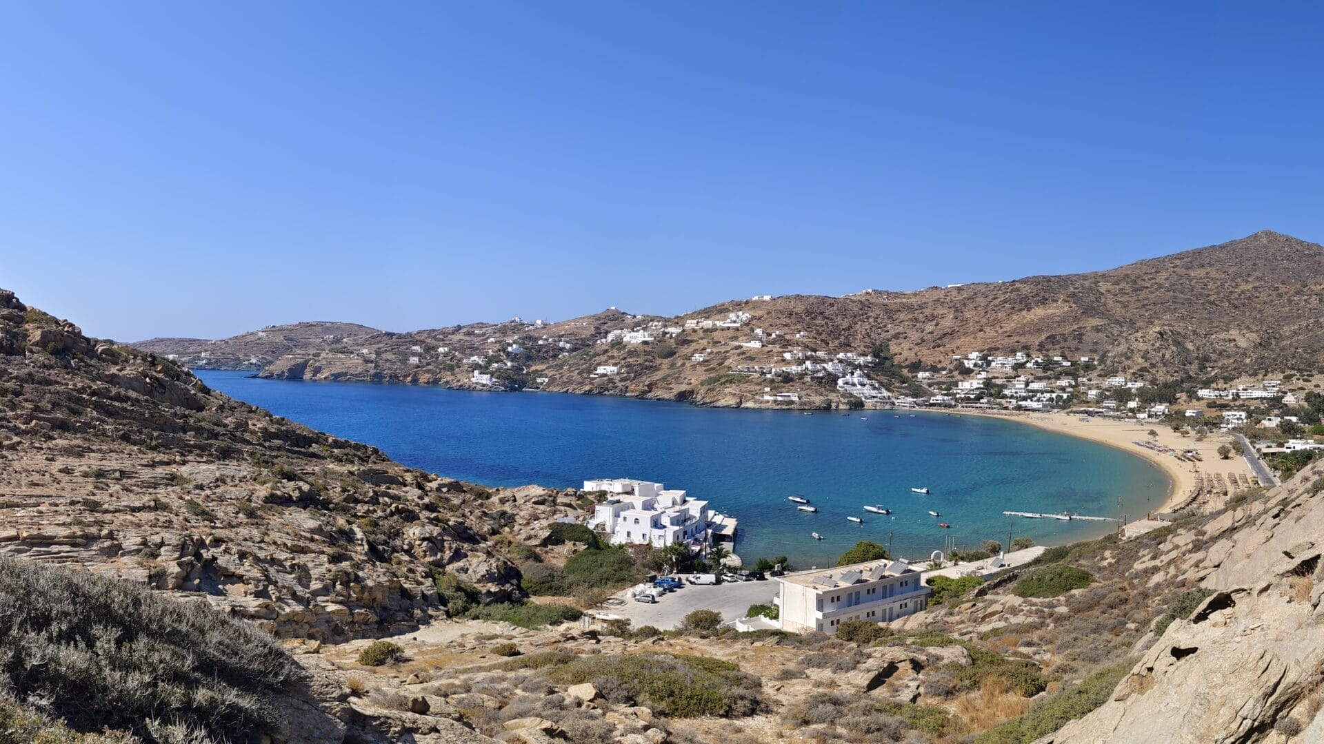 Eine Küstenlandschaft mit Sandstrand, klarem blauen Wasser und verstreuten weißen Gebäuden vor felsigen Hügeln unter klarem Himmel. Boote liegen in Küstennähe vor Anker.