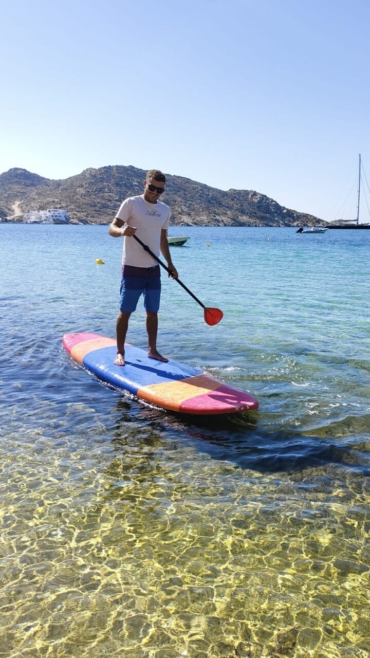 Eine Person paddelt auf klarem, seichtem Wasser in der Nähe einer felsigen Küste unter einem strahlenden Himmel.