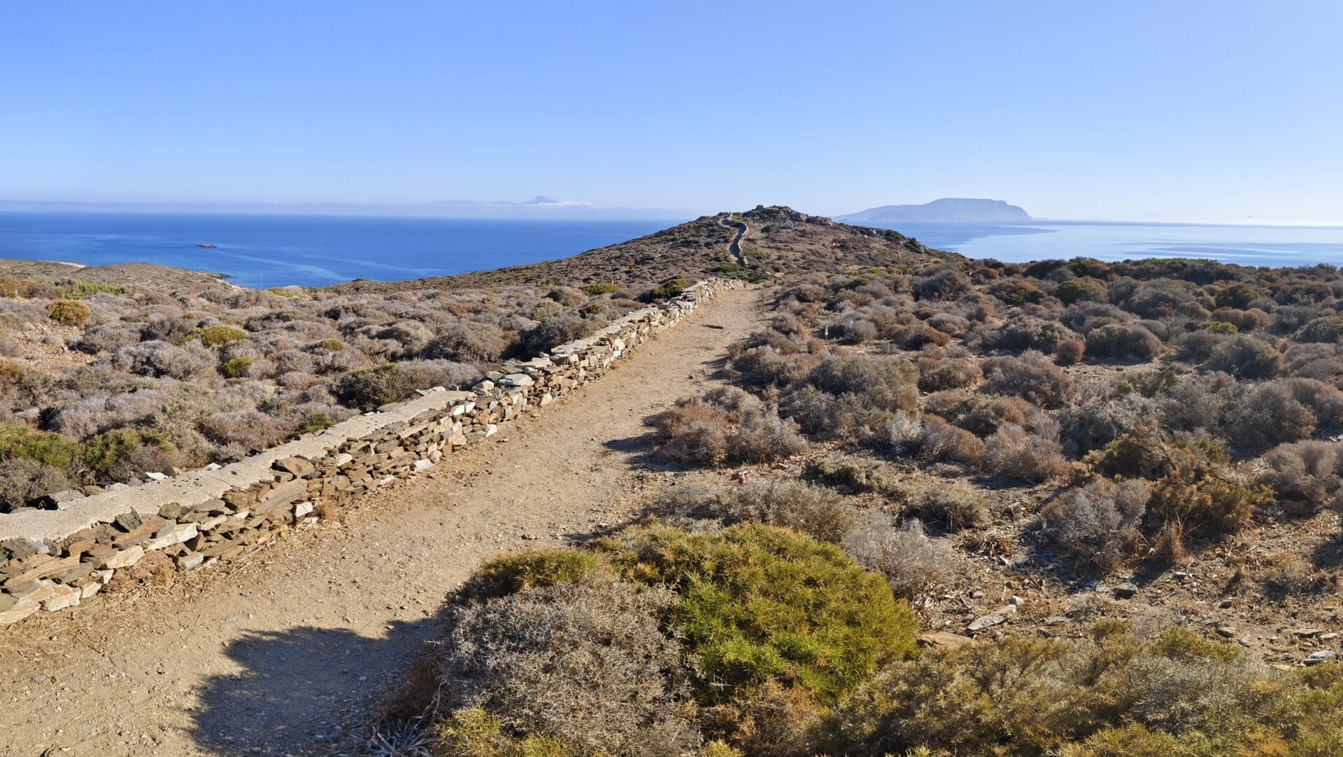 Ein steiniger Pfad führt durch trockene Vegetation zum Meer. Unter einem klaren blauen Himmel sind in der Ferne Inseln zu sehen.