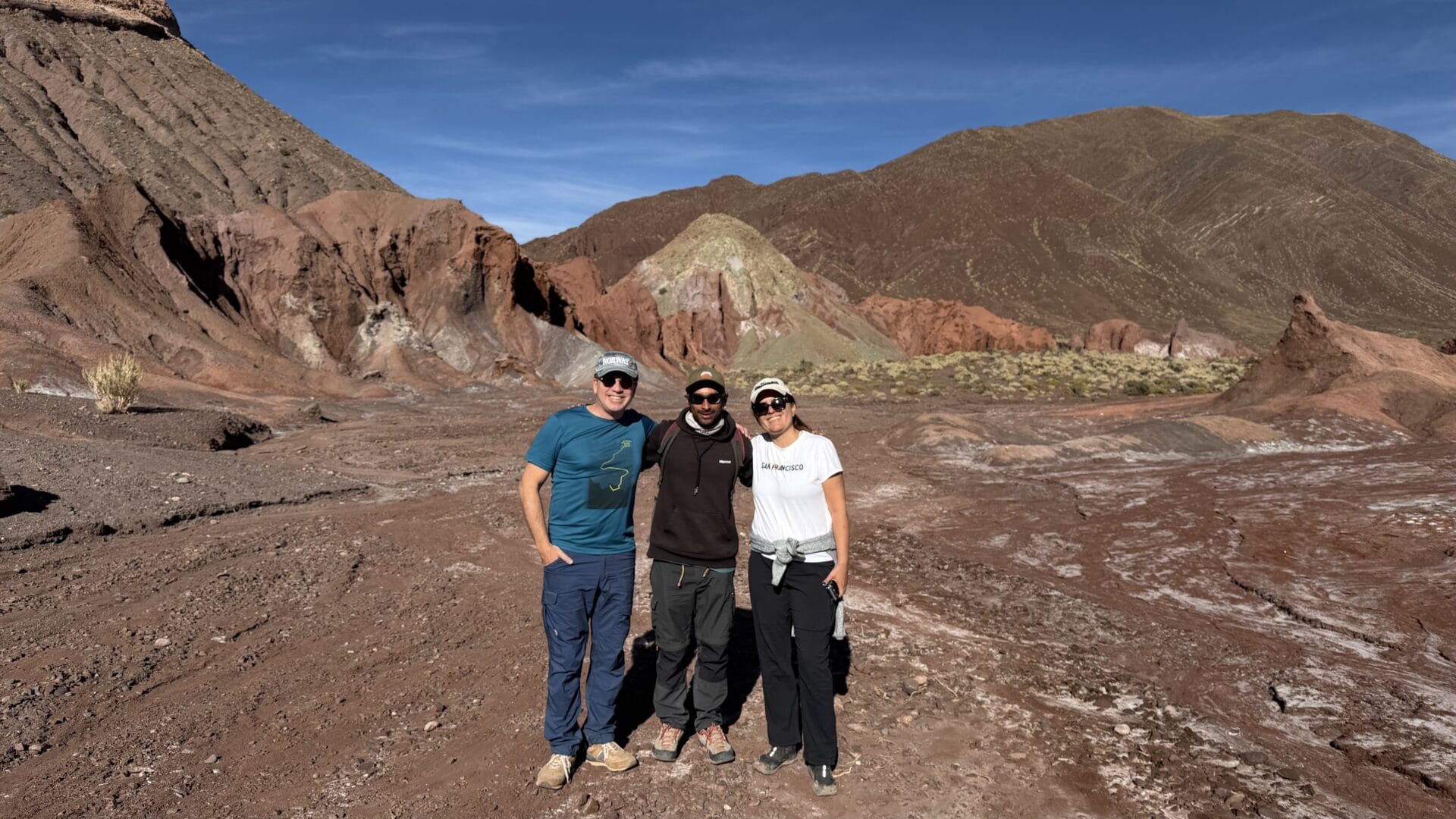 Drei Menschen stehen zusammen auf felsigem Gelände in der Atacama-Wüste Chiles, mit bunten Hügeln und einem klaren blauen Himmel im Hintergrund.
