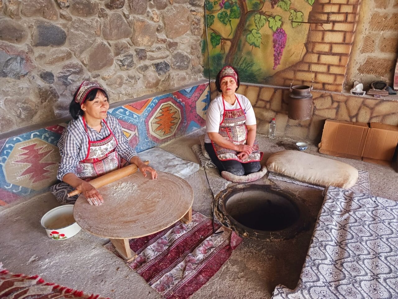 Zwei Frauen in traditioneller Kleidung bereiten an einem runden Tisch und einem Tandoor-Ofen auf Teppichen sitzend in einem mit Wandmalereien verzierten Steinraum Teig zu.
