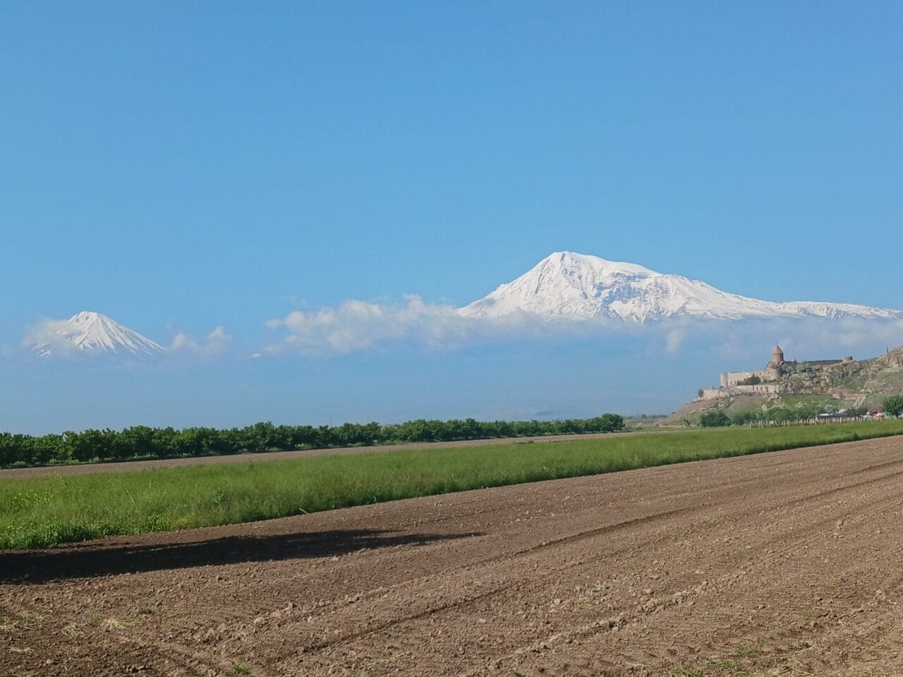 In der Ferne erheben sich schneebedeckte Berge hinter einem grünen Feld, gepflügtem Boden und einem kleinen Kloster oder einer Festung am rechten Bildrand.