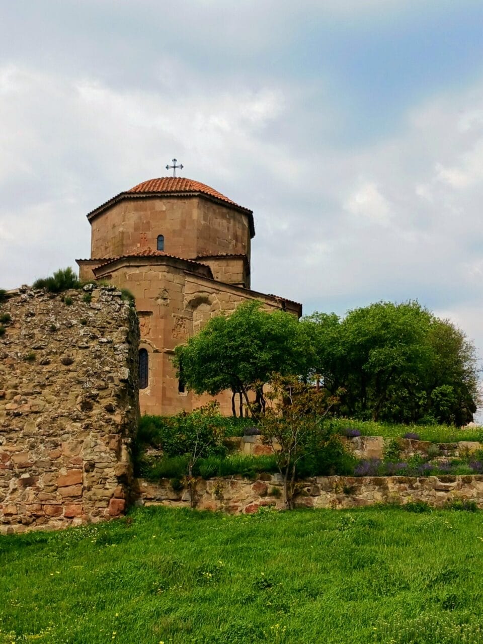 Eine historische Steinkirche mit einer rot gekachelten Kuppel steht hinter alten Steinmauern und grünen Bäumen unter einem teilweise bewölkten Himmel.