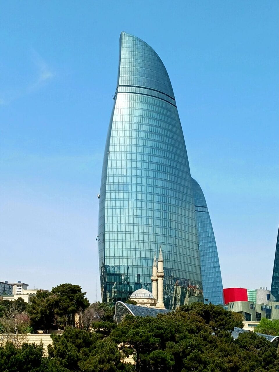 Ein hoher, moderner Wolkenkratzer aus Glas erhebt sich über den Bäumen, an dessen Fuß eine kleine historische Moschee mit Minaretten zu sehen ist, vor dem Hintergrund eines klaren blauen Himmels.