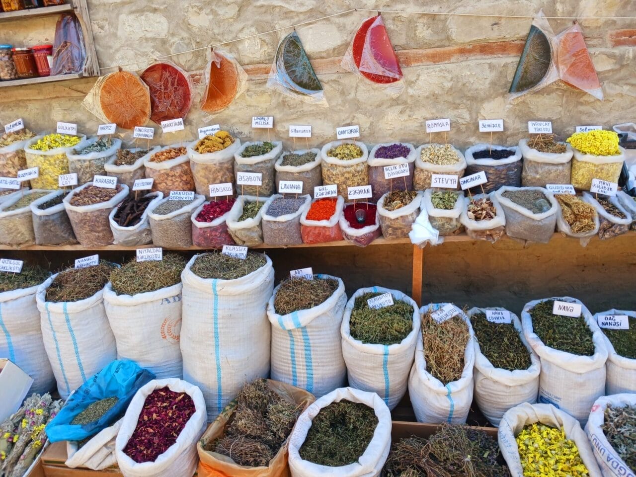 Eine Vielzahl von getrockneten Kräutern und Gewürzen, die in offenen Säcken und Beuteln auf einem Markt im Freien ausgestellt werden, jedes mit seinem Namen vor einer Steinmauer beschriftet.