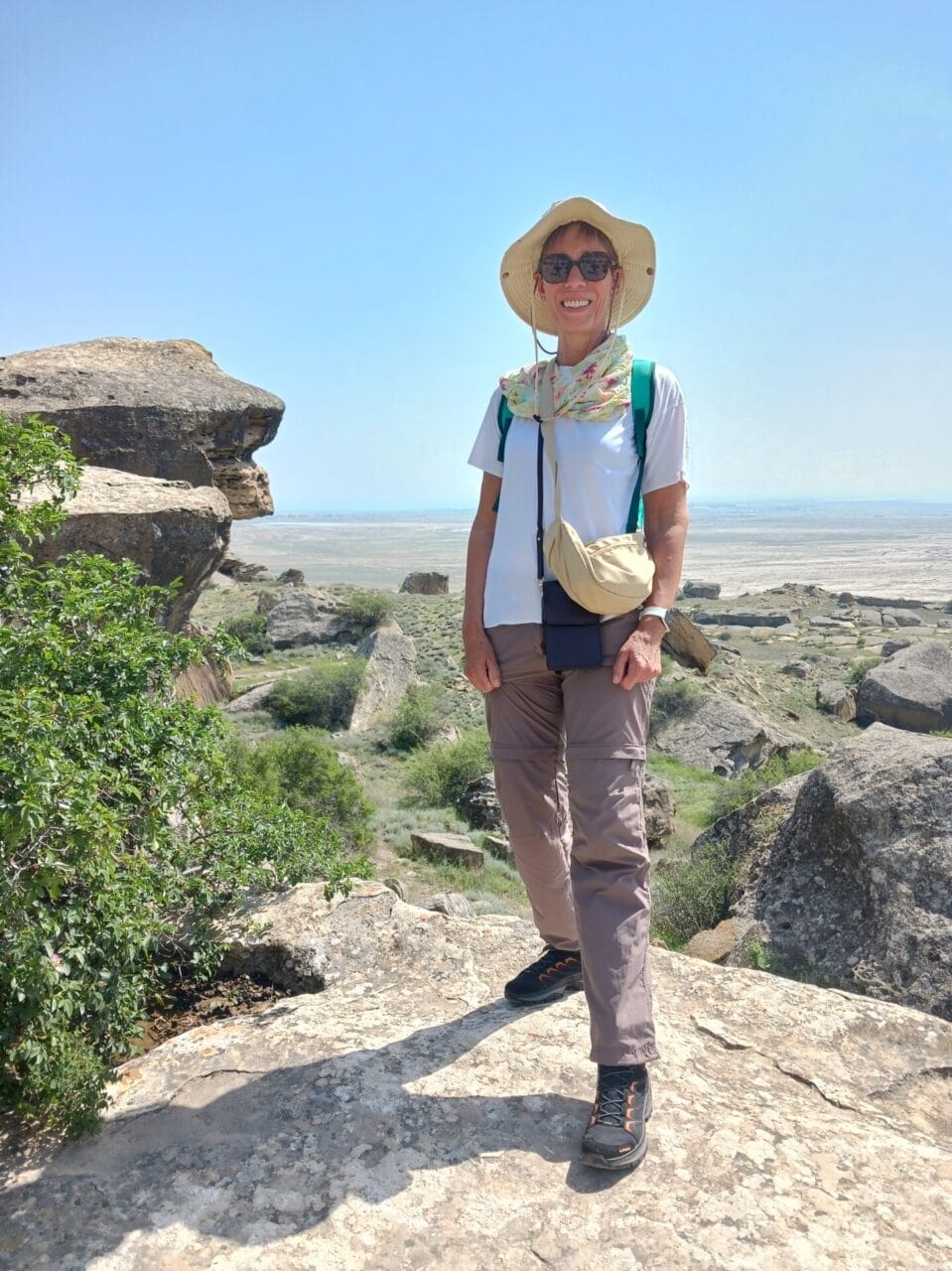 Eine Frau mit Sonnenhut, Sonnenbrille und Wanderkleidung steht auf einer felsigen Landschaft mit viel Grün und einer weiten Ebene im Hintergrund unter einem klaren Himmel.