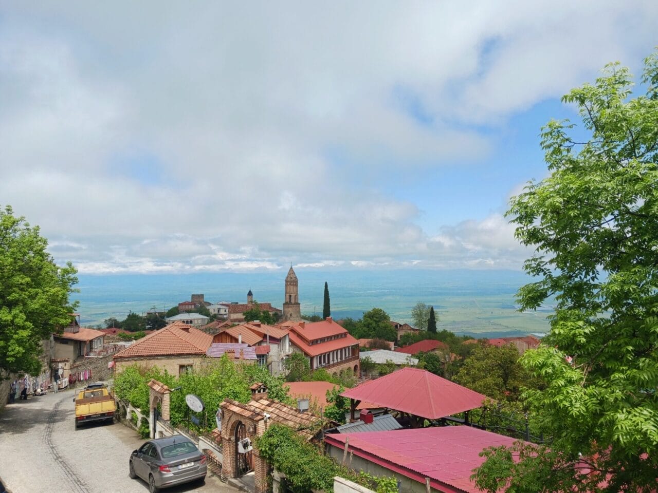 Eine Stadt am Hang mit roten Dächern, einem Kirchturm, Autos auf einer kurvenreichen Straße und einem weiten Landschaftsausblick unter einem teilweise bewölkten Himmel.