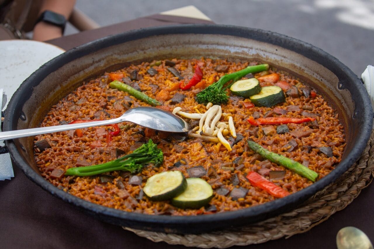 Albufera-Paella Eine Pfanne mit Gemüsepaella mit Reis, Zucchini, Brokkoli, roter Paprika, Spargel und einem Löffel, der auf dem Rand liegt.