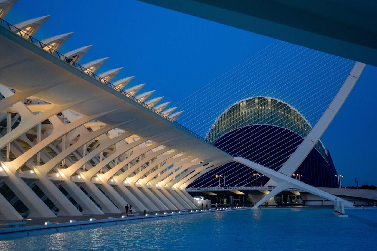 Valencia - L'Hemisferic Moderne architektonische Strukturen mit kantigen weißen Balken und einem großen Kuppelbau neben einem Wasserbecken, fotografiert in der Abenddämmerung.