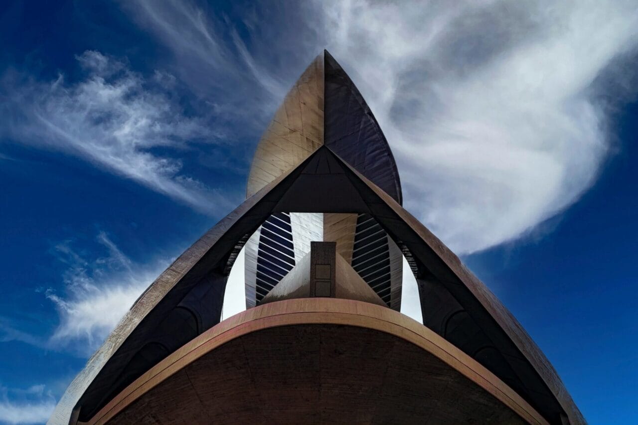Valencia - Stadt der Künste Modernes Gebäude, von unten fotografiert, mit geschwungenen architektonischen Linien und einem spitzen Dach vor einem blauen Himmel mit Wolkenfetzen.