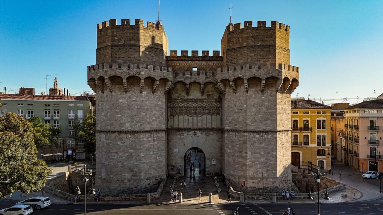 Torres de Serranos Großes mittelalterliches Steintor mit zwei zylindrischen Türmen und Zinnen, umgeben von städtischen Gebäuden und ein paar Autos und Fußgängern, unter einem klaren blauen Himmel.