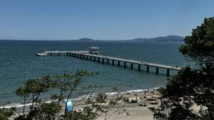 Ein langer Pier erstreckt sich von einem Sandstrand mit Menschen, Sonnenschirmen und Bäumen im Vordergrund über das Meer; in der Ferne sind Hügel zu sehen.