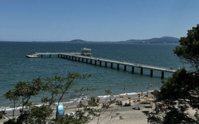 Bulgarien neu entdecken – Sweti Vlas zwischen Sonne, Stil und Lebensfreude Ein langer Pier erstreckt sich von einem Sandstrand mit Menschen, Sonnenschirmen und Bäumen im Vordergrund über das Meer; in der Ferne sind Hügel zu sehen.