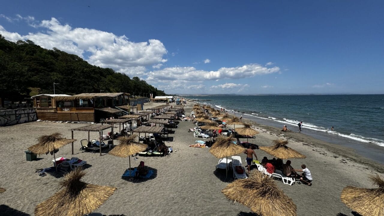 Burgas Strand-Promenade Strand mit Strohschirmen und Liegestühlen, Menschen, die sich entspannen und schwimmen, Holzkonstruktionen auf der linken Seite, ein bewaldeter Hügel und ein teilweise bewölkter Himmel.