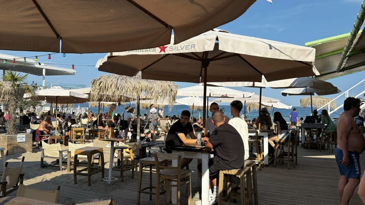 In einem belebten Strandcafé sitzen Menschen an Tischen unter großen Sonnenschirmen, im Hintergrund ist das Meer zu sehen, und über dem Kopf scheint die Sonne.