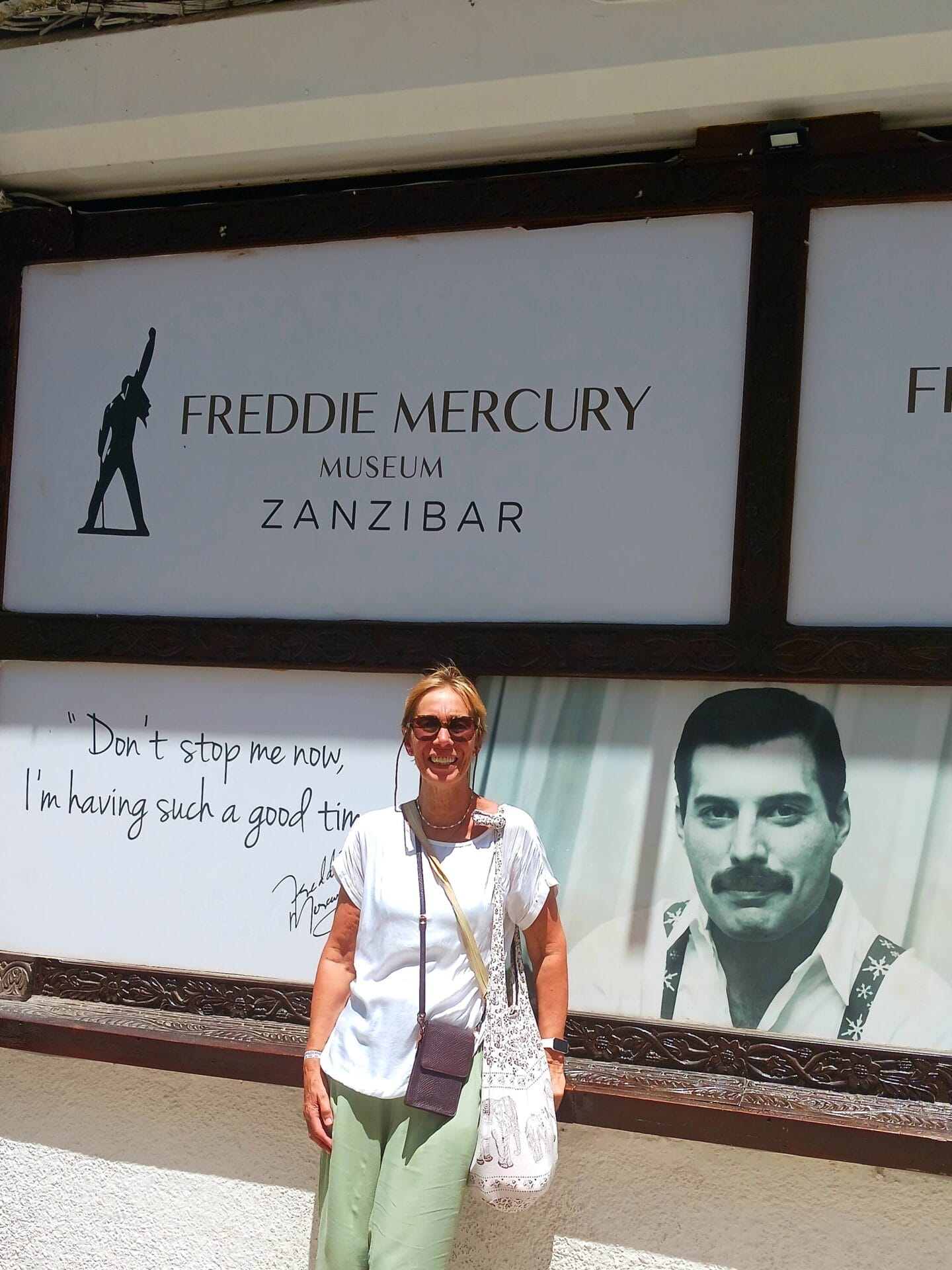 Eine Frau steht lächelnd vor dem Schild des Freddie-Mercury-Museums in Sansibar. An der Wand hinter ihr hängen ein großes Foto von Freddie Mercury und ein Zitat.