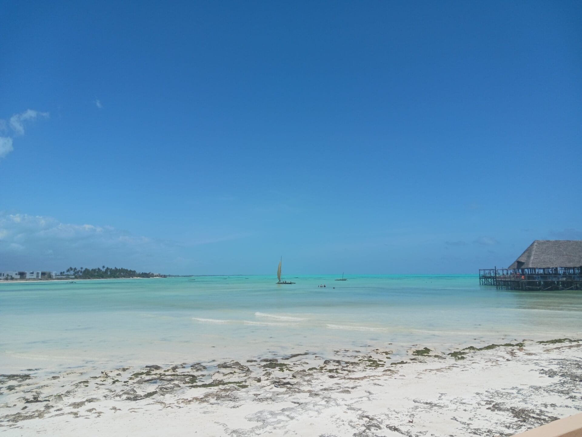 Ein Sandstrand mit Seegras führt zu türkisfarbenem, flachem Wasser. Zwei kleine Segelboote sind auf dem Wasser, und auf der rechten Seite steht eine Holzkonstruktion auf Stelzen unter einem klaren blauen Himmel.