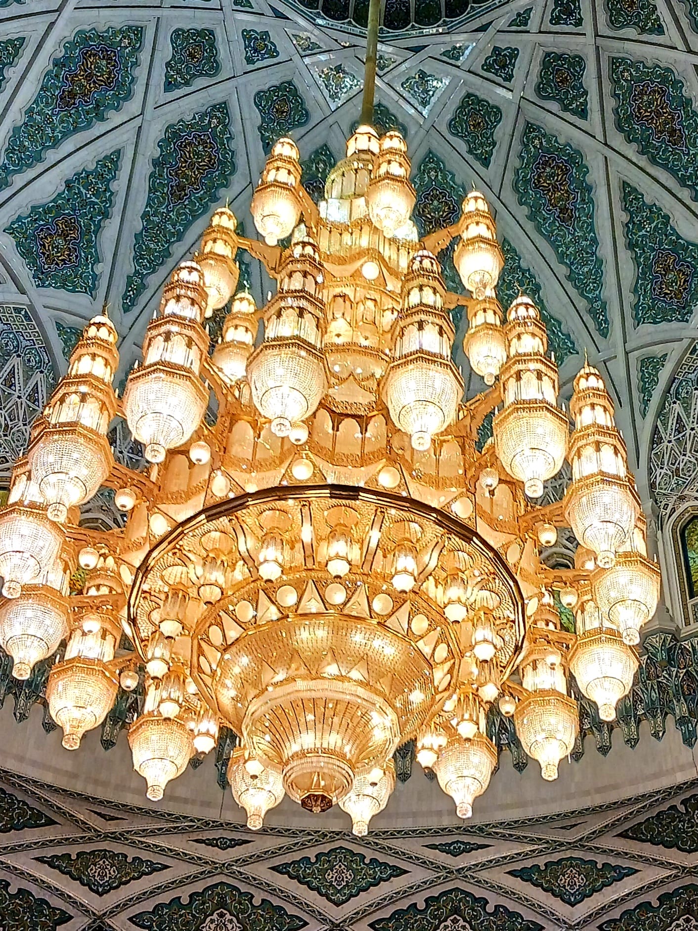 Ein großer, verschnörkelter Kristalllüster mit mehreren Ebenen hängt von einer dekorativen, gemusterten Kuppeldecke.