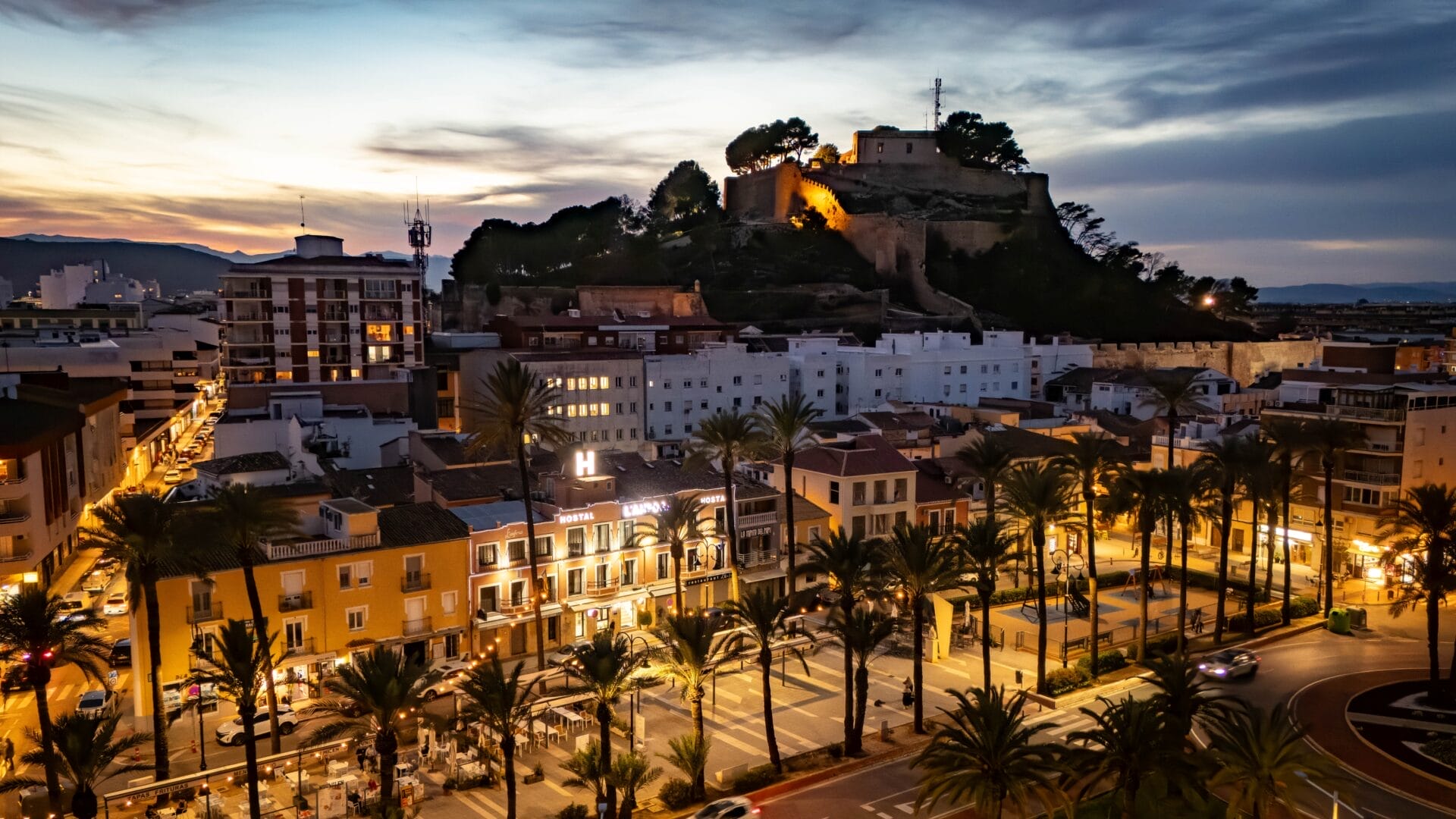Ein Stadtbild in der Abenddämmerung zeigt eine Festung auf einem Hügel mit Blick auf beleuchtete Gebäude, Palmen und Straßen im Vordergrund.