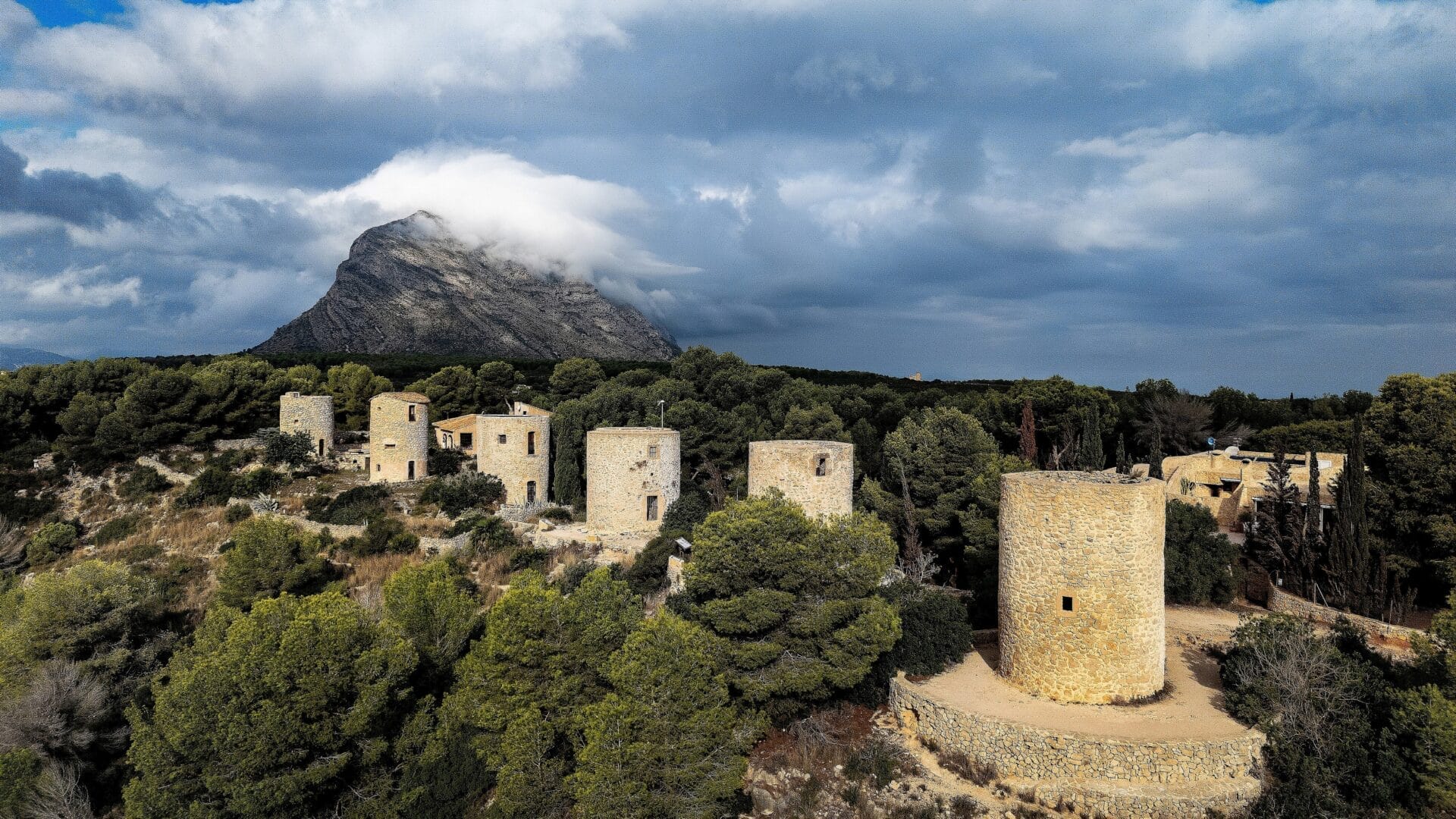 Eine Reihe alter steinerner Windmühlen steht auf einem von Bäumen umgebenen Hügel, mit einem großen, teilweise von Wolken bedeckten Berg im Hintergrund.