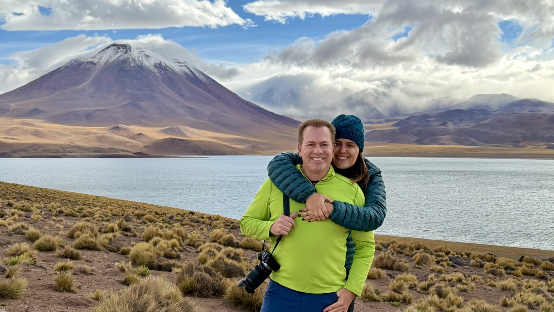 Zwei Personen stehen lächelnd an einem See in der bergigen Landschaft von Piedras Rojas, wobei die eine die andere von hinten umarmt, eingerahmt von schneebedeckten Gipfeln und treibenden Wolken im Hintergrund.