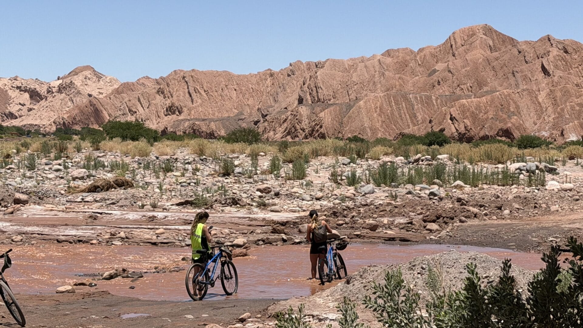Zwei Personen mit Fahrrädern stehen in der Nähe eines seichten, schlammigen Baches in einer felsigen Wüstenlandschaft mit rötlichen Bergen im Hintergrund.